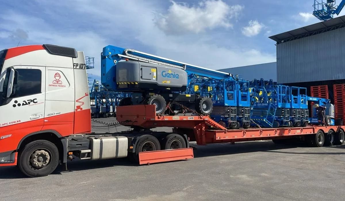 Carreta do Grupo APC carregando uma plataforma Genie no pátio da empresa, com a frota ao fundo