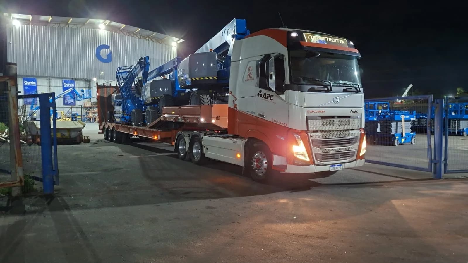 Caminhão da APC com prancha carregada de plataformas elevatórias Genie, saindo para entrega.